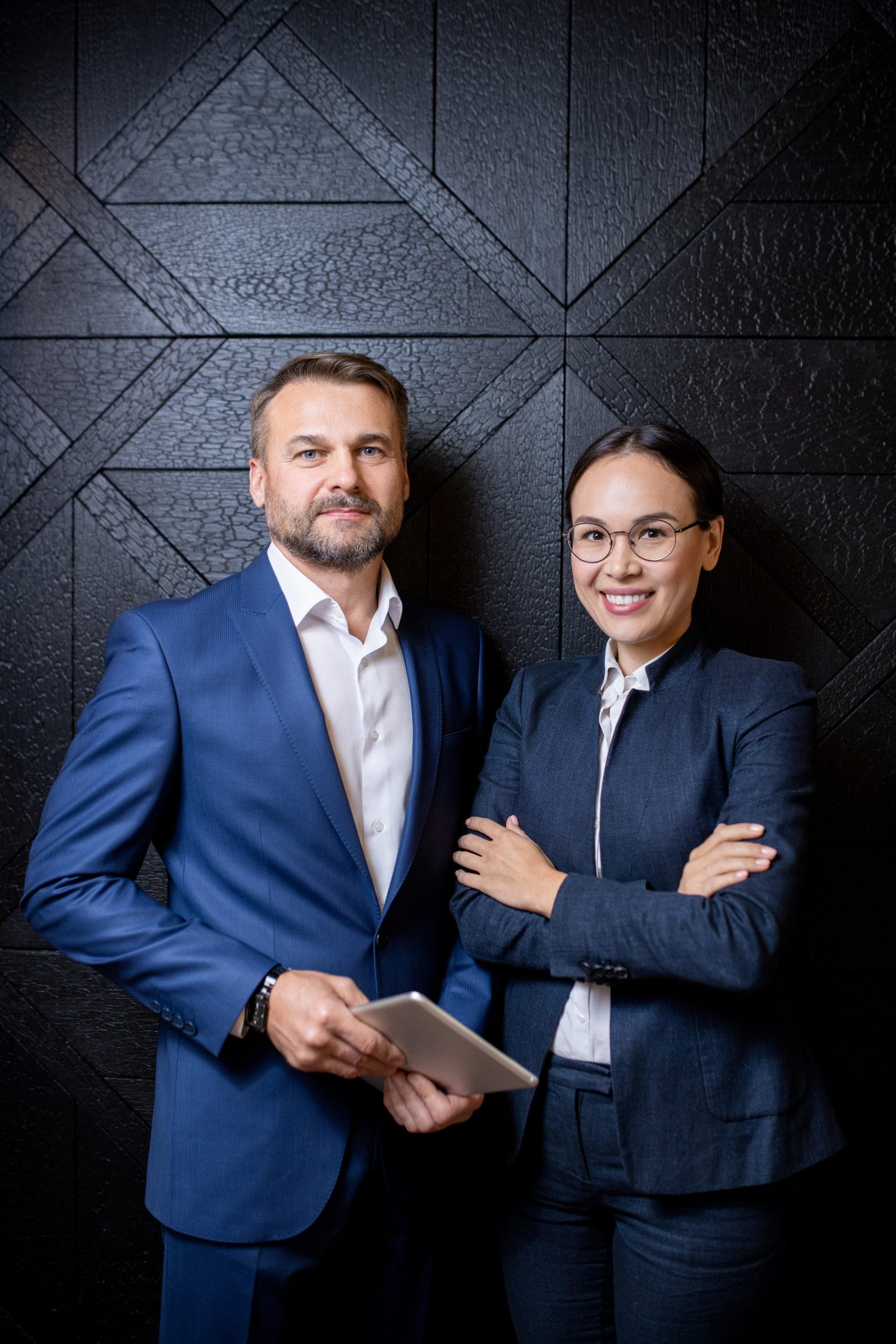 Happy young elegant businesswoman and her middle aged confident colleague with touchpad standing by black wall